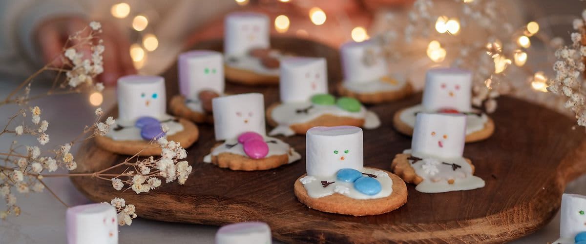 Dítě pozoruje sušenky s marshmallows a barevnými bonbony na dřevěném prkénku, ozdobené světýlky.