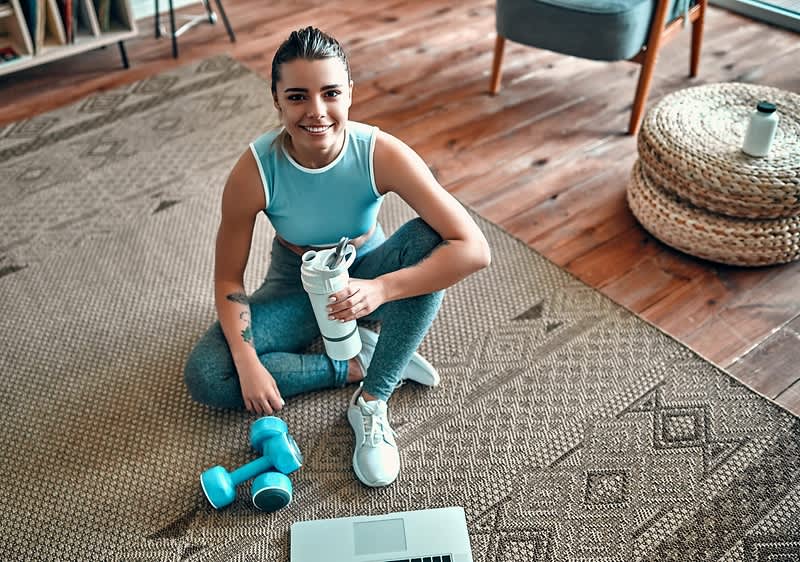 Žena v sportovním oblečení sedí na koberci s činkou a lahví vody, před ní je otevřený notebook.
