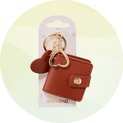 Rotes Mini-Portemonnaie aus Kunstleder mit Druckknopf, an einem goldfarbenen Schlüsselring mit goldenem Herzanhänger und kleinem roten Herzanhänger befestigt, auf weißer Pappkarte mit der Aufschrift „AMAZING“