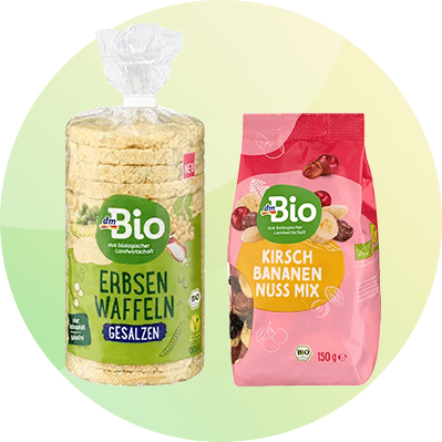 Zwei dmBio-Produkte vor hellem Kreis hintergrund: links transparente Packung mit runden Erbsen-Waffeln, grünes Etikett „dmBio ERBSEN WAFFELN GESALZEN“, rechts rosa Standbeutel „dmBio KIRSCH BANANEN NUSS MIX“ 150 g