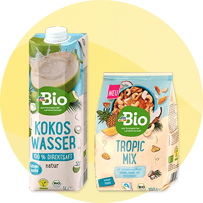 Zwei dmBio-Produkte vor hellgelbem Hintergrund: links eine 1‑L-TetraPak-Packung Kokoswasser mit Aufschrift „Kokos Wasser 100% Direktsaft natur“, rechts eine Tüte „Tropic Mix“ mit Nüssen und Früchten und dmBio-Logo