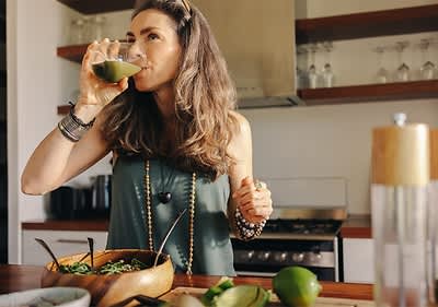 Frau in Küche trinkt aus Glas mit Limetten und Salat auf Tisch