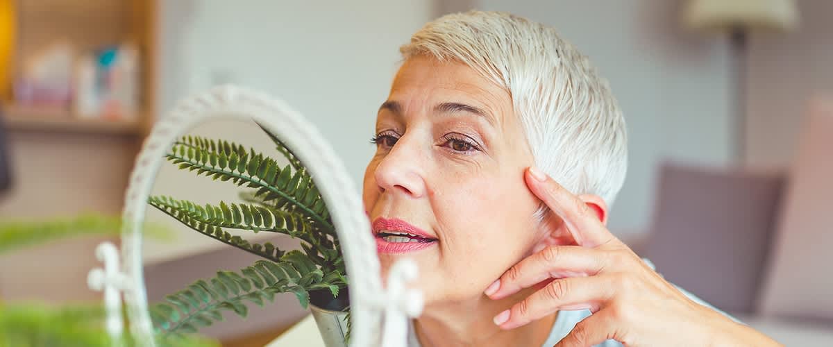 Aeltere Frau betrachtet ihr Gesicht im Kosmetikspiegel