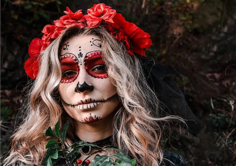Frau mit weiß geschminktem Calavera-Gesicht, roten Augenringen und schwarzen Verzierungen, trägt roten Blumenkranz, lange helle Locken, schwarzes Tüllnetz im Haar und hält grüne Pflanzenblätter vor dem Oberkörper