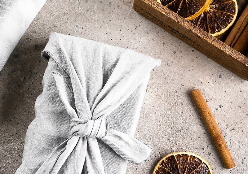 Ein Geschenk in hellgrauem Stoff mit Knoten liegt auf hellem Tisch, daneben getrocknete Orangenscheiben, eine Zimtstange und eine Holzkiste mit weiteren getrockneten Orangenscheiben