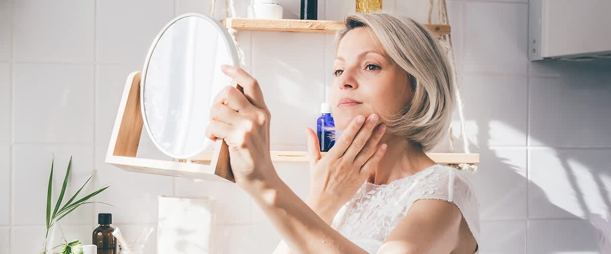 Frau mit blonden Schulterlangen Haaren betrachtet ihr Gesicht im Kosmetikspiegel