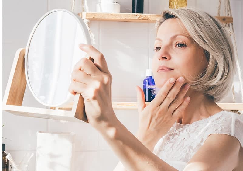 Frau mit blonden Schulterlangen Haaren betrachtet ihr Gesicht im Kosmetikspiegel