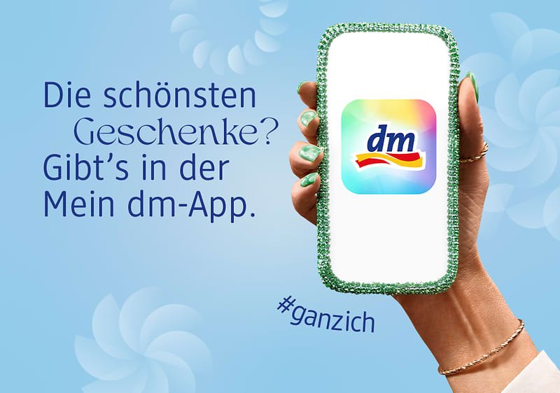 Eine Hand hält ein Smartphone mit dem dm-Logo, daneben der Text „Die schönsten Geschenke? Gibt’s in der Mein dm-App“ und „#ganzich“