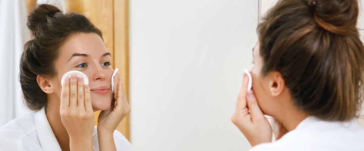 Frau bei der Gesichtsreinigung vor dem Spiegel mit jeweils einem Wattepads auf jeder Wange