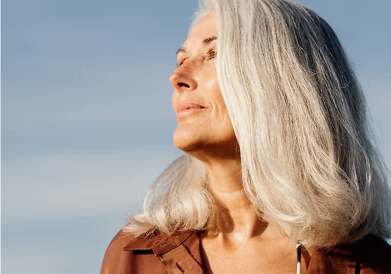 Eine Frau mit grauem Haar trägt eine braune Bluse vor einem blauen Himmel.