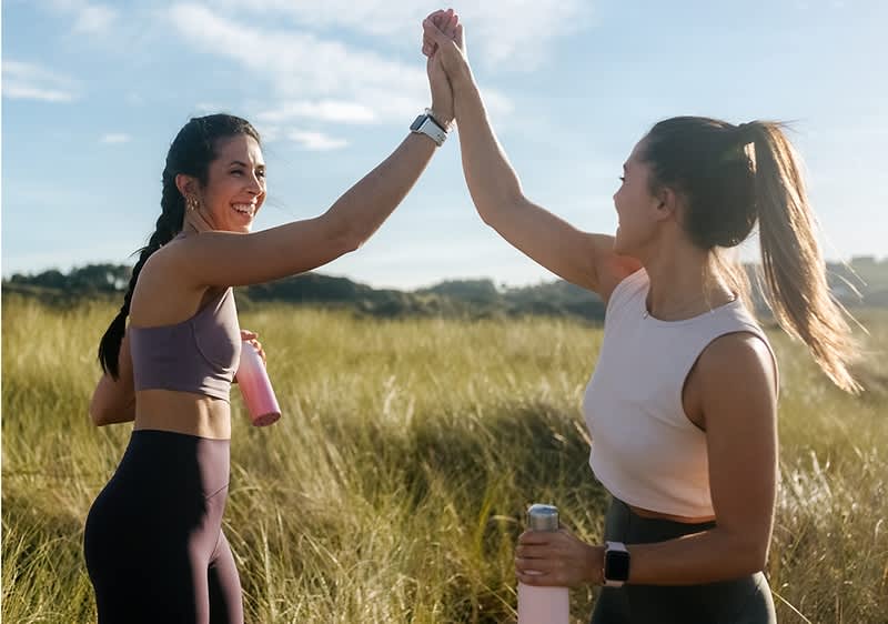 Zwei Frauen in Sportkleidung geben sich im Freien ein High-Five, beide halten eine rosa Wasserflasche in der Hand.