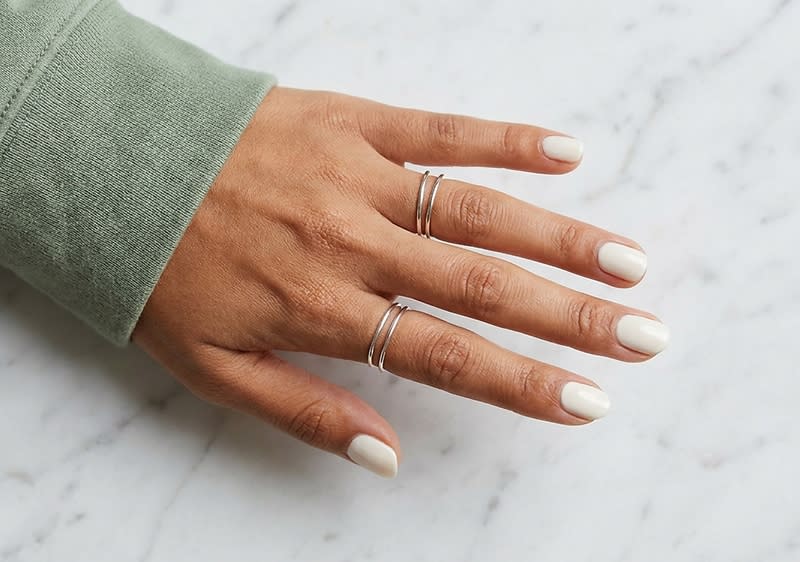 Rechte Hand mit hellem, cremeweiß lackierten Nägeln, Manschette eines grünen Pullovers sichtbar, an Mittelfinger und Ringfinger schmale silberne Stapelringe auf marmorierter Oberfläche
