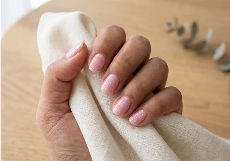 Hand mit gepflegten, kurz gefeilten Nägeln in zartem rosa Glitzerlack, die ein helles Leinenstück hält