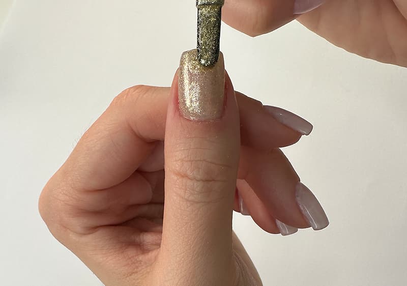 Daumen mit frisch lackiertem, goldglitzerndem Nagel, darüber wird mit einem Pinsel goldener Nagellack aufgetragen
