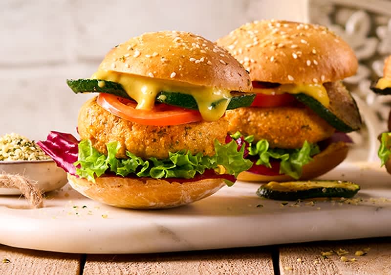 Zwei Sesambrötchen-Burger mit knuspriger Gemüse-Patty, Tomatenscheiben, grünen Salatblättern und schmelzendem Käse, auf einem weißen Brett angerichtet