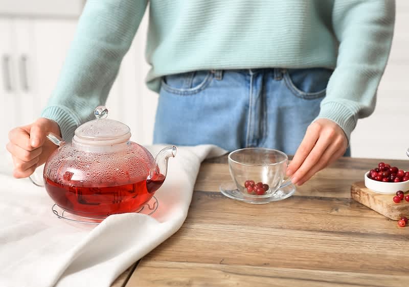 Teezubereitung mit rotem Früchtetee und Beeren in einer Glaskanne und Tasse auf Holztisch