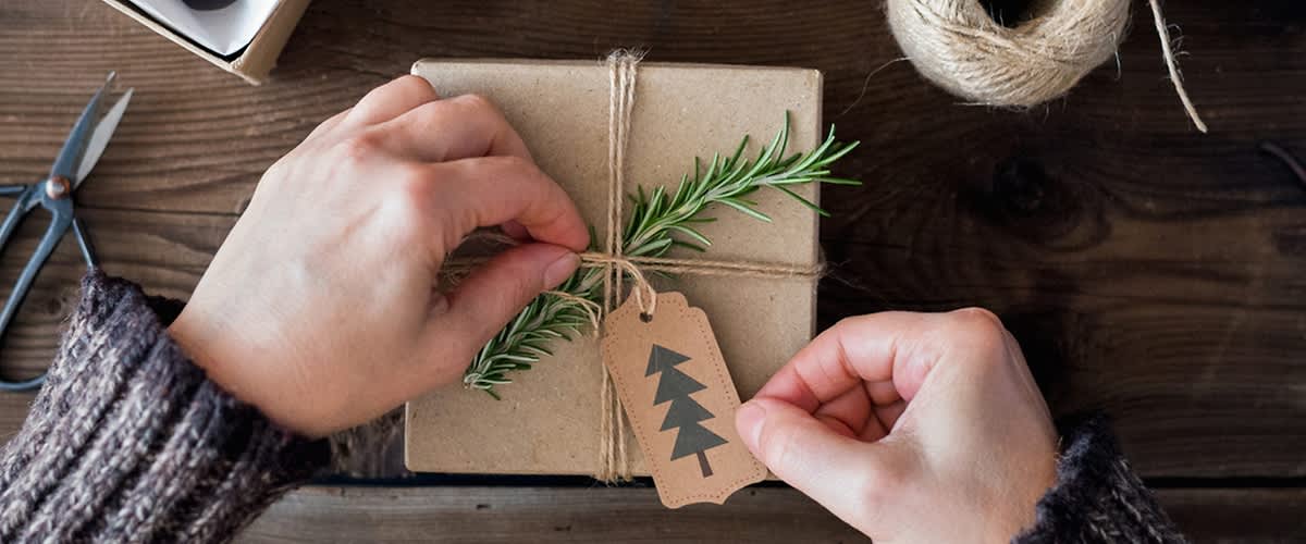 Zwei Hände binden ein Geschenk mit braunem Papier und einer Kordel, dekoriert mit einem Rosmarinzweig und einem Etikett mit einem Tannenbaum-Symbol