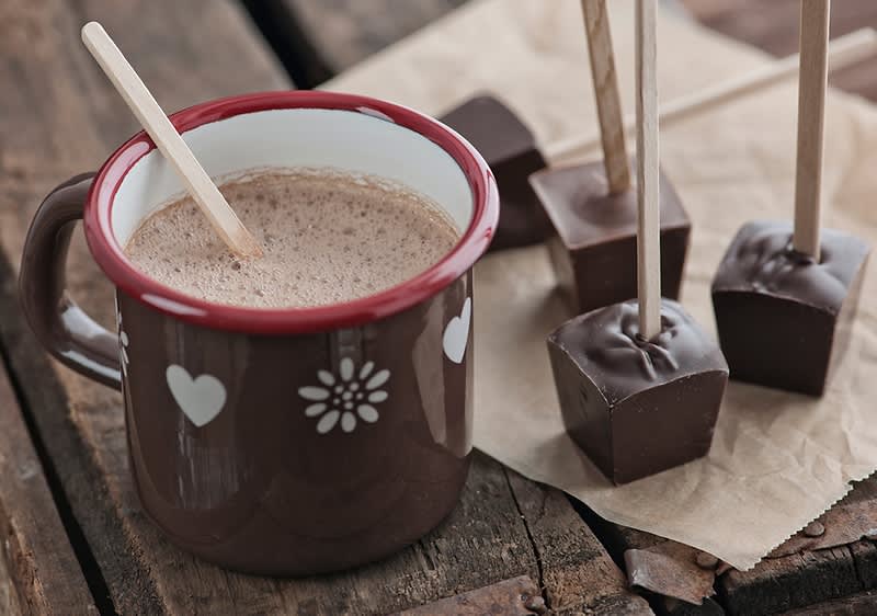 Ein brauner Keramikbecher mit weißen Herz- und Blumenmotiven, gefüllt mit heißer Schokolade, steht auf einem Holztisch neben mehreren Schokoladenstücken auf Holzstäbchen