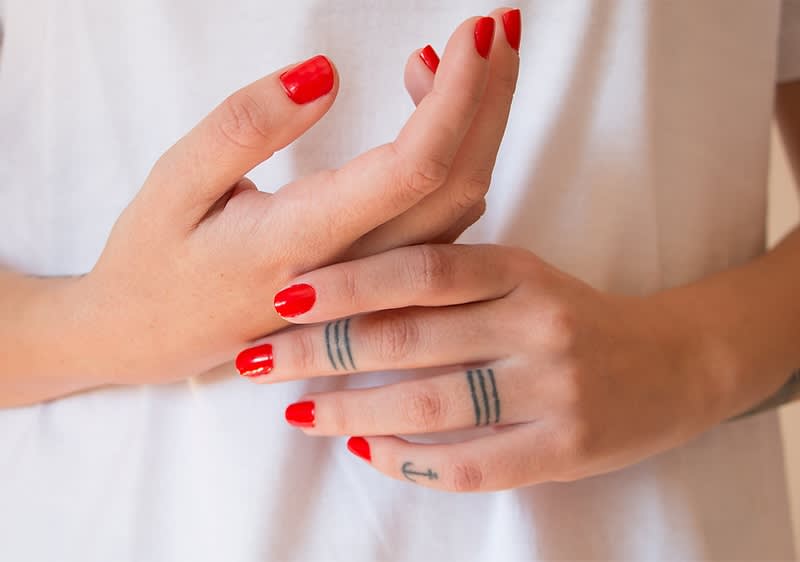Rote Fingernägel und Tattoos auf Händen vor weißem Shirt