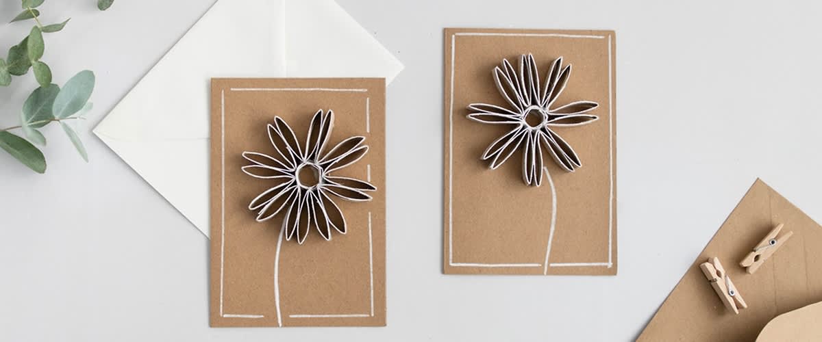 Zwei braune Karten mit weißen Blumen aus Papierstreifen, daneben ein weißer Umschlag.