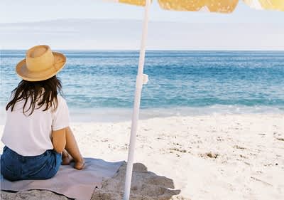 Sommerlich gekleidete Frau mit Sonnenhut am Strand unter Sonnenschirm