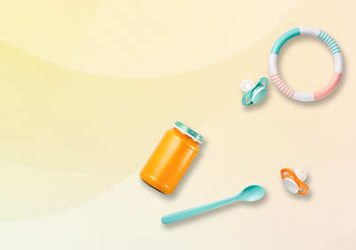 Ein Glas mit orangefarbenem Inhalt, ein türkisfarbener Löffel, zwei Schnuller und ein buntes Beißring-Spielzeug auf hellem Hintergrund