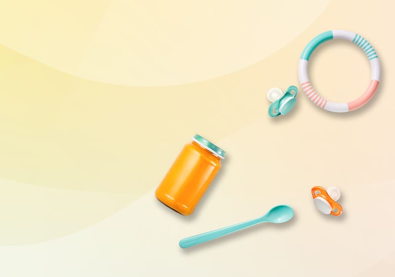 Ein Glas mit orangefarbenem Inhalt, ein türkisfarbener Löffel, zwei Schnuller und ein buntes Beißring-Spielzeug auf hellem Hintergrund