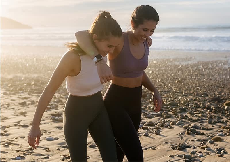 Zwei Personen in Sportkleidung gehen am Strand entlang und haben die Arme umeinander gelegt. Im Hintergrund sind Meer und Kieselsteine zu sehen.