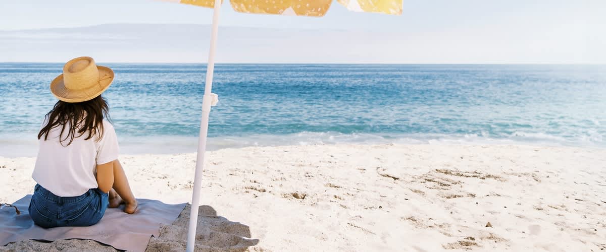 Sommerlich gekleidete Frau mit Sonnenhut am Strand unter Sonnenschirm