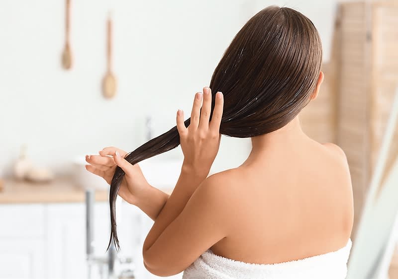 Eine Frau mit langen, glatten Haaren steht mit dem Rücken zur Kamera und hält ihr Haar in der Hand, während sie ein Handtuch um den Körper trägt