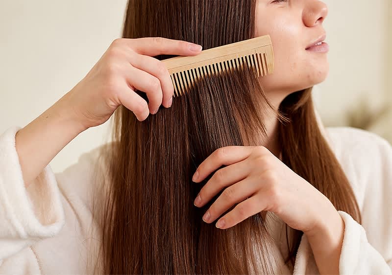Eine Person in einem weißen Bademantel kämmt ihr langes, glattes braunes Haar mit einem Holzkamm