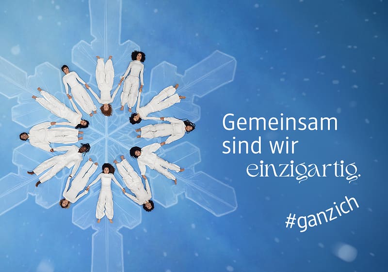 Acht Personen in weißen Anzügen liegen kreisförmig auf einem blauen Hintergrund mit Schneeflockenmuster, daneben der Text „Gemeinsam sind wir einzigartig“ und „#ganzich“