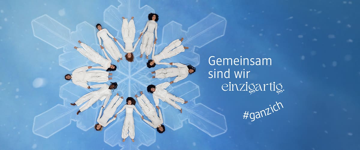 Acht Personen in weißen Anzügen liegen kreisförmig auf einem blauen Hintergrund mit Schneeflockenmuster, daneben der Text „Gemeinsam sind wir einzigartig“ und „#ganzich“