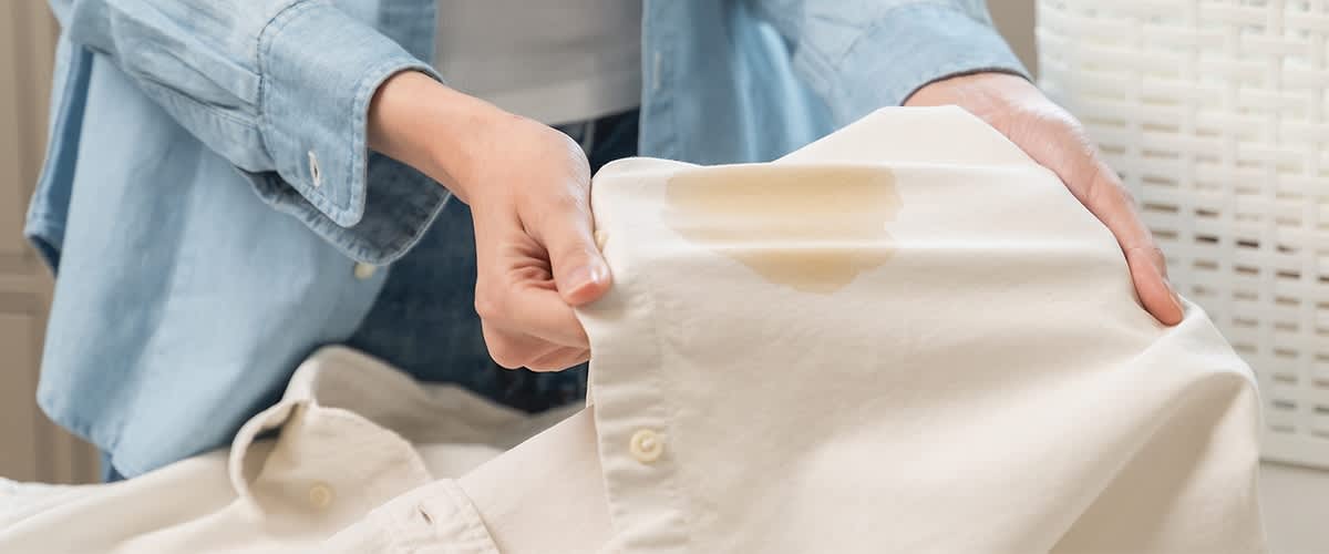 Eine Person hält ein weißes Hemd mit einem großen, hellbraunen Fleck auf der Vorderseite.