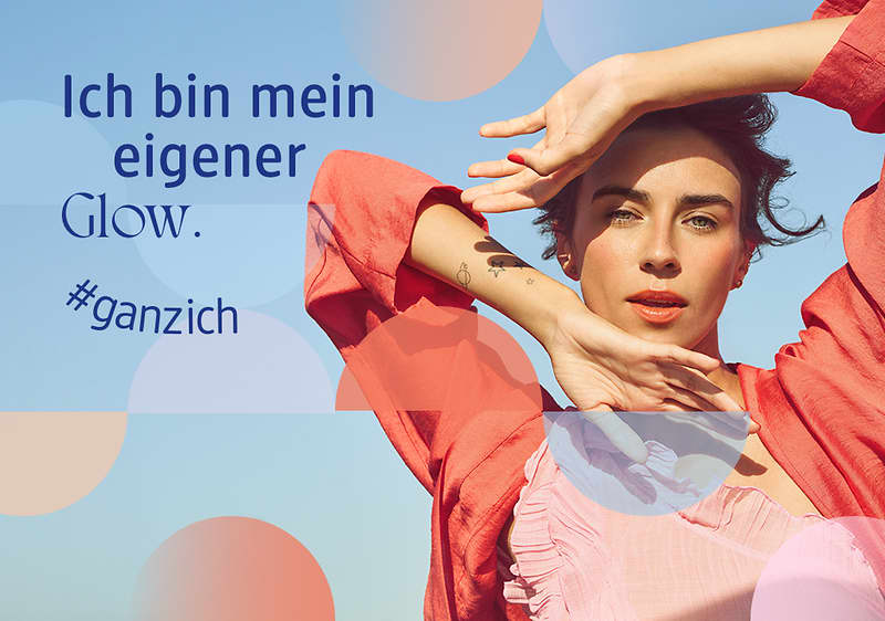 Frau in rotem Oberteil mit erhobenen Armen vor blauem Himmel, links blauer Text „Ich bin mein eigener Glow. #ganzich“