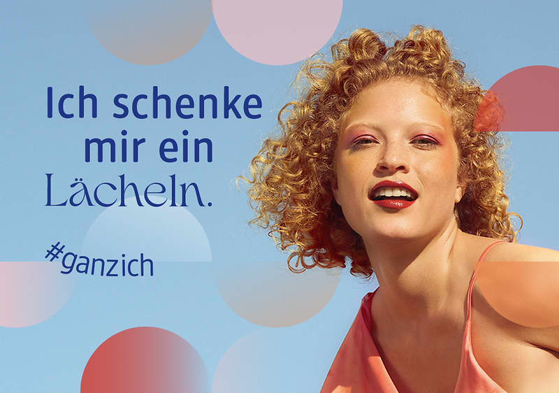 Frau mit lockigem Haar in einem orangenen Top vor blauem Hintergrund mit farbigen Kreisen, links Text „Ich schenke mir ein Lächeln.“ und Hashtag „#ganzich“