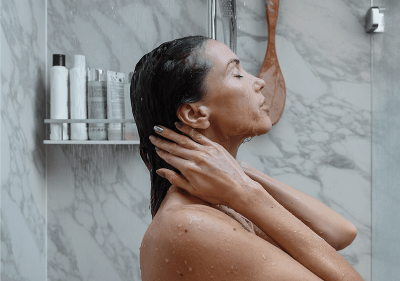 Frau unter Dusche mit Shampoo-Flaschen im Hintergrund