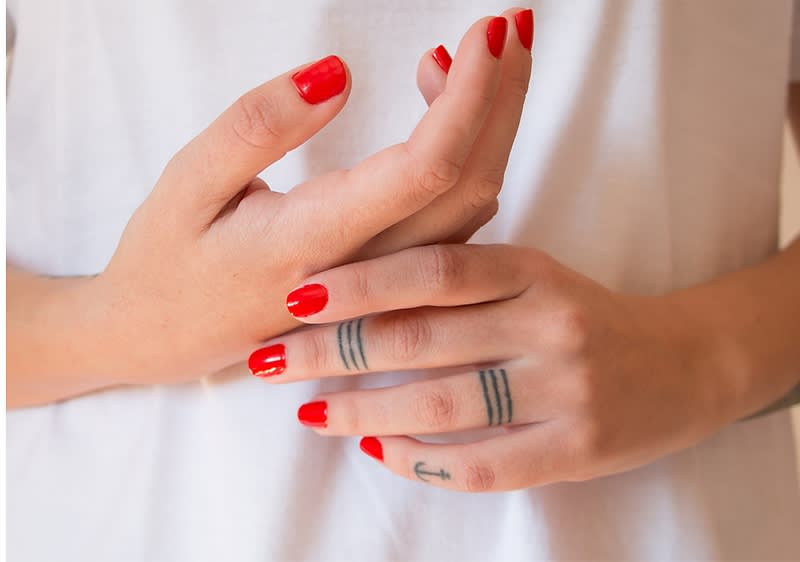 Rote Fingernägel und Tattoos auf Händen vor weißem Shirt