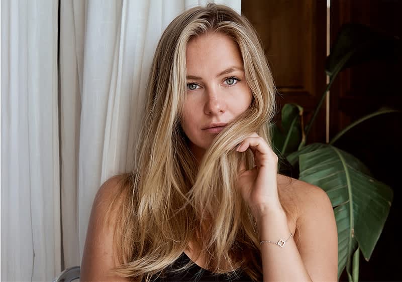 Blonde Frau mit langen Haaren vor Vorhang und Pflanze