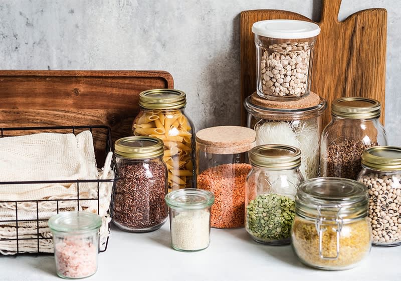 Vorratsgläser mit verschiedenen Lebensmitteln auf Holzbrett und Korb