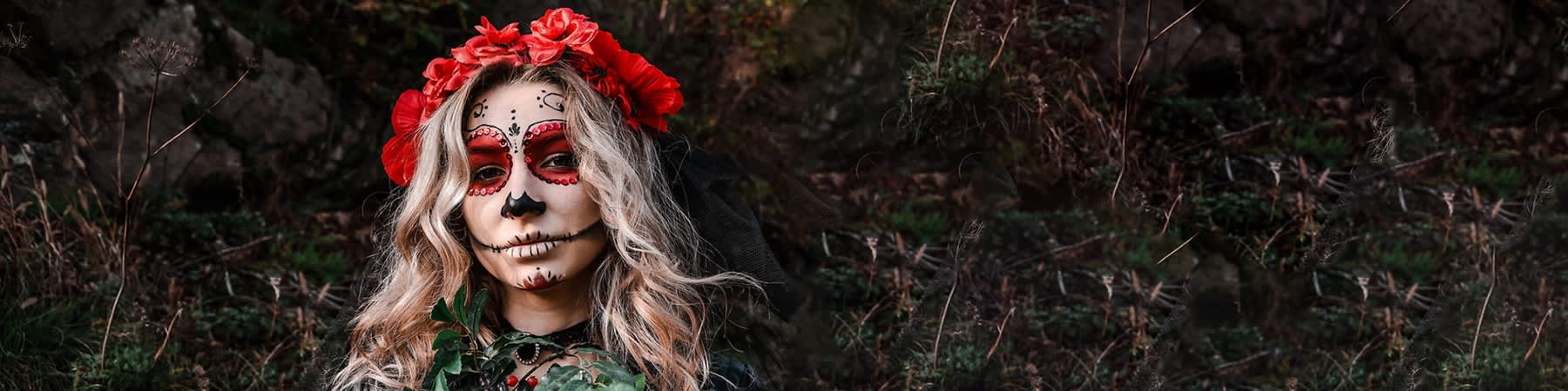 Frau mit Totenkopf-Schminke im Día-de-los-Muertos-Stil, rote Augenringe und schwarze Nasen- und Mundlinien, blonder lockiger Haare, roter Blumenkranz und schwarzer Schleier, trägt grüne Blätter im Vordergrund