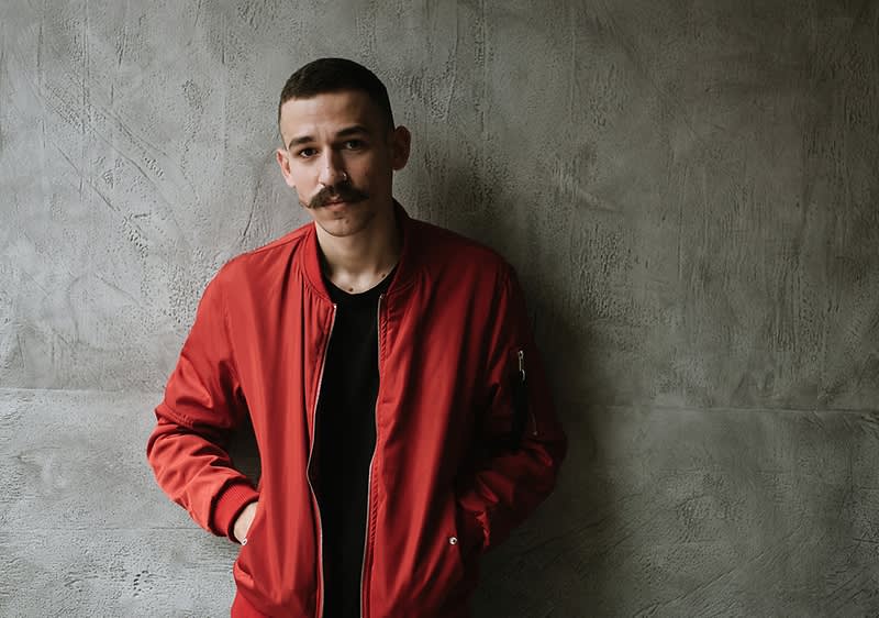 Ein Mann mit Schnurrbart und roter Jacke und vor grauer Wand