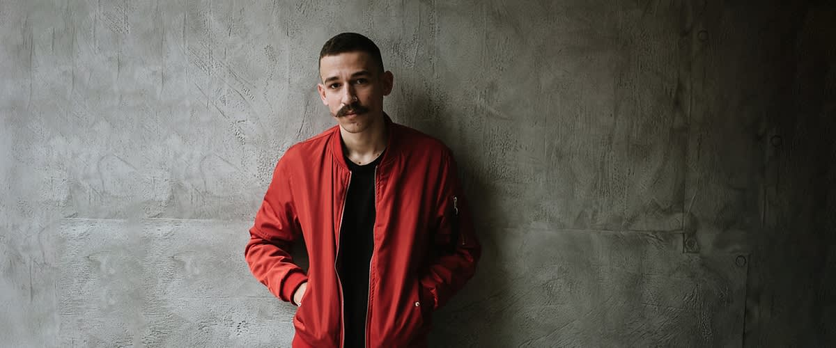 Ein Mann mit Schnurrbart und roter Jacke und vor grauer Wand