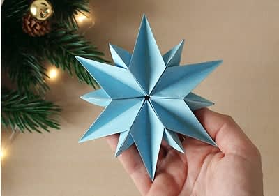 Eine Hand hält einen blauen, gefalteten Origami-Stern, im Hintergrund sind Tannenzweige und eine kleine Lichterkette sichtbar