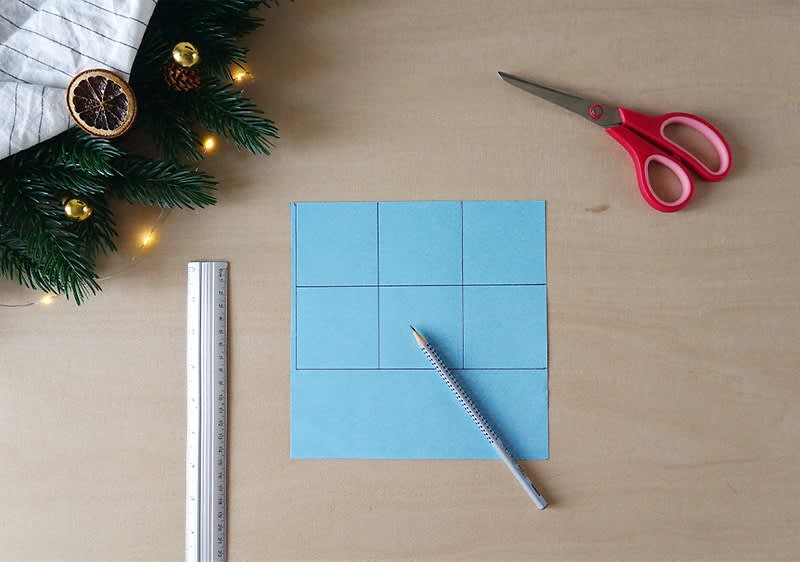 Ein blauer Papierbogen mit einem 3x3 Raster liegt auf einem Holztisch, daneben ein silberner Stift, eine rote Schere und ein Lineal, links oben Tannenzweige mit Lichtern und einer getrockneten Orangenscheibe