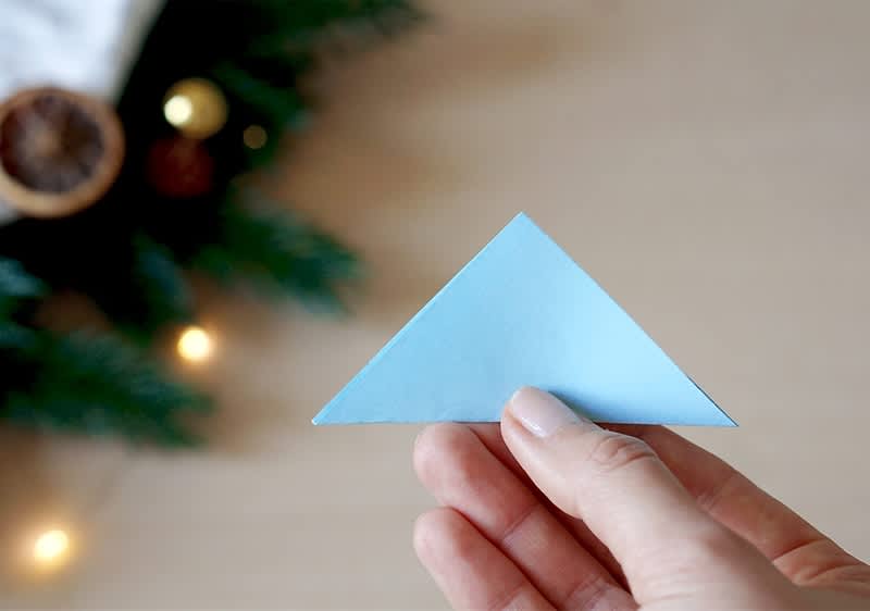Eine Hand hält ein blaues Papierdreieck, im Hintergrund sind unscharfe Tannenzweige und Lichter zu sehen