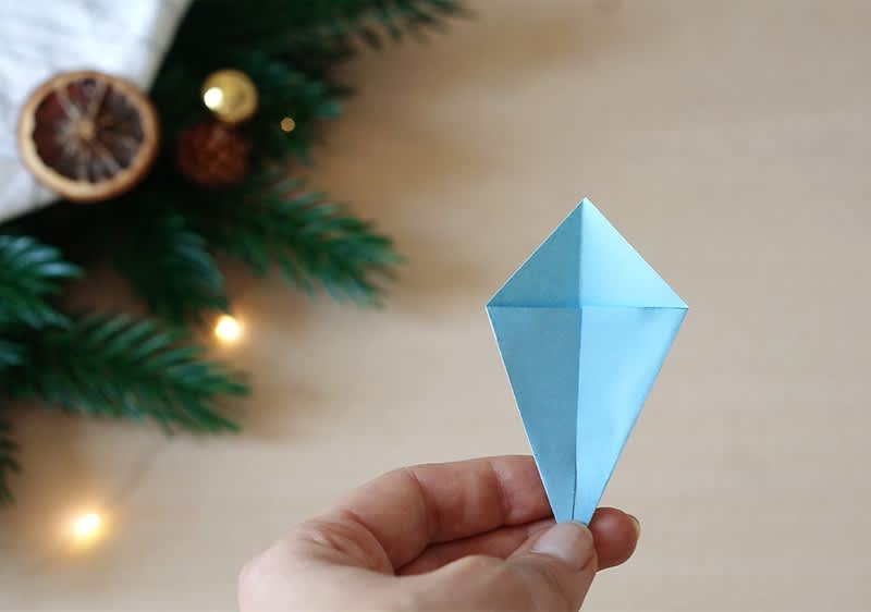 Eine Hand hält einen blauen Origami-Diamanten aus Papier, im Hintergrund sind Tannenzweige und eine getrocknete Orangenscheibe mit Lichtern sichtbar