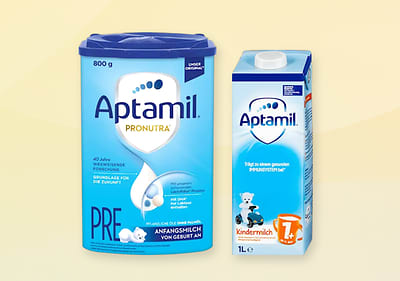 Eine blaue Aptamil PRE Packung mit Bärchenmotiv und eine weiße Aptamil Kindermilch Packung mit Bärchenmotiv und orangefarbener Eins