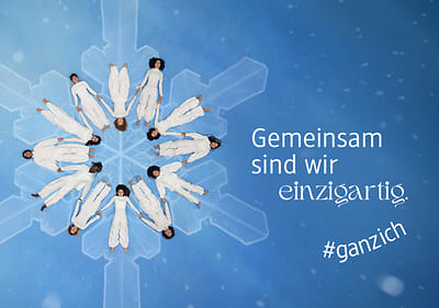 Acht Personen in weißen Anzügen liegen kreisförmig auf einem blauen Hintergrund mit Schneeflockenmuster, daneben der Text „Gemeinsam sind wir einzigartig“ und „#ganzich“