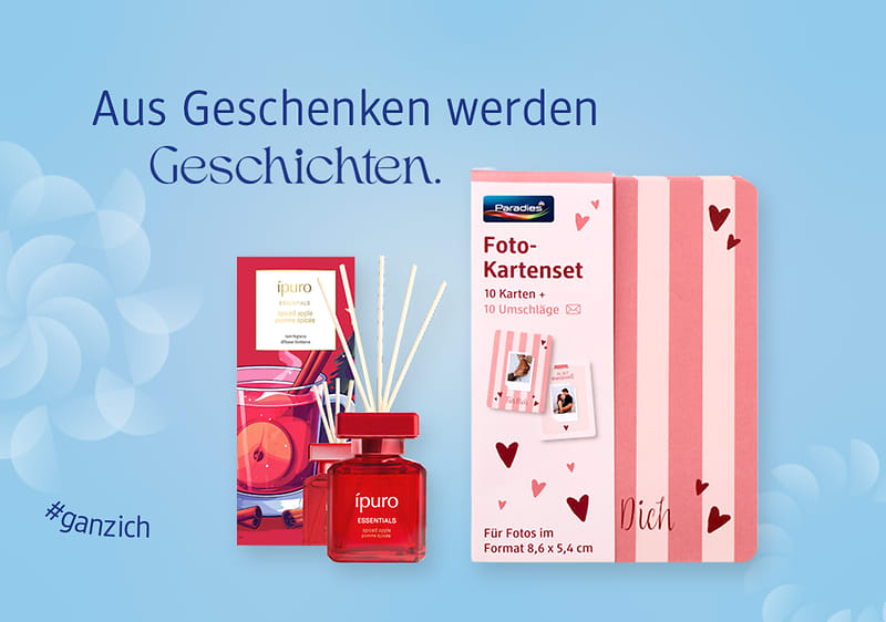 Auf hellblauem Hintergrund links ein rotes Raumduftset mit Flakon und Rattanstäbchen, rechts eine rosa-gestreifte Foto-Kartenbox mit Aufschrift „Foto-Kartenet“ und Herzmotiven, oben Text „Aus Geschenken werden Geschichten“